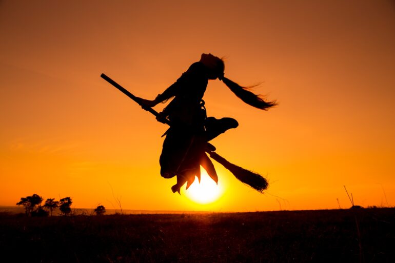 Silhouette einer jungen Frau, die mit einem Besen zwischen den Beinen vor der untergehenden Sonne in die Luft springt. Es sieht aus, als würde sie auf dem Besen fliegen.