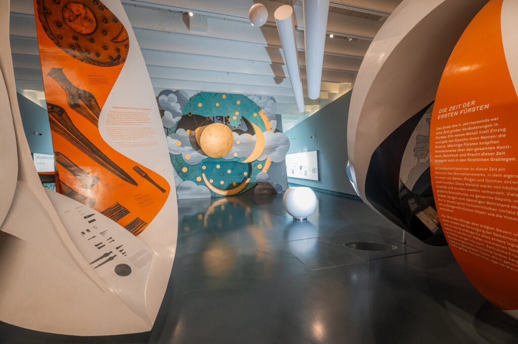Weiträumiger Ausstellungsbereich der Arche Nebra mit orangefarbenen und weißen geschwungenen Ausstellungselementen, Infotafeln zur Bronzezeit und Blick auf die Himmelsscheibeninstallation. 