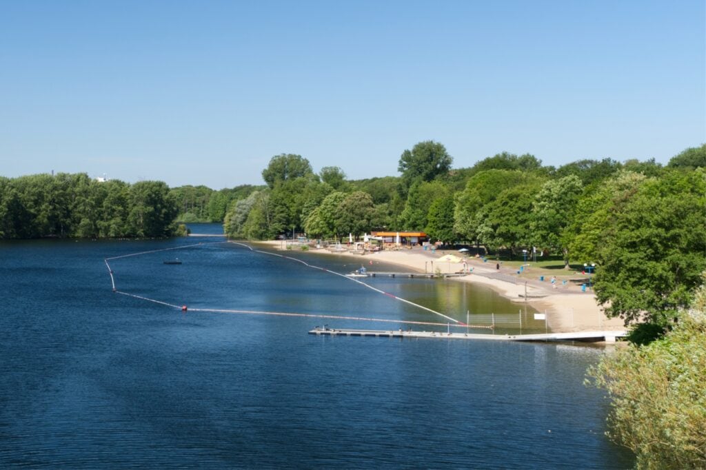 Ein langer Sandstrand an einem Waldsee, an dem mehrere Menschen sich sonnen.