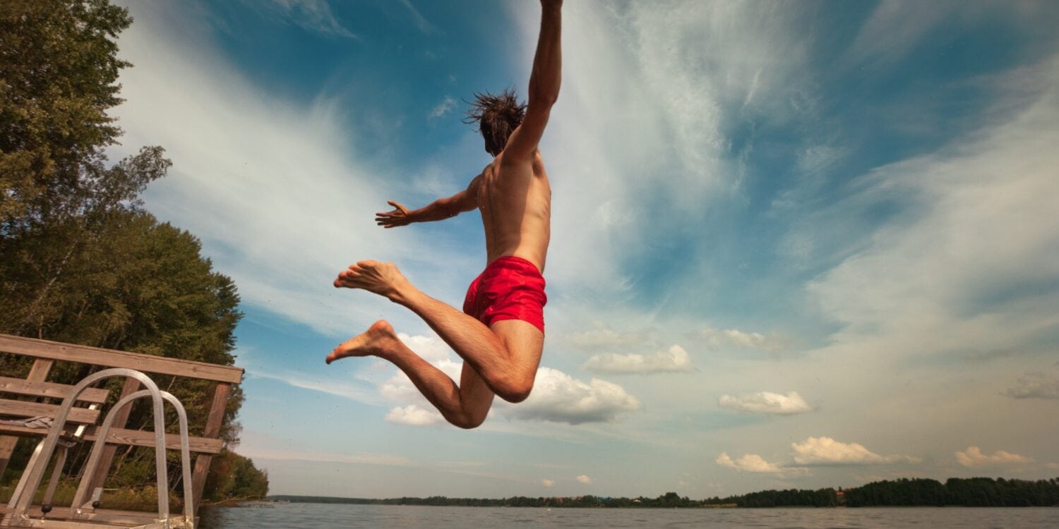 Ein Mann in roter Badehose springt mit ausgebreiteten von einem kleinen Steg in einen See.