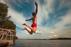 Ein Mann in roter Badehose springt mit ausgebreiteten von einem kleinen Steg in einen See.