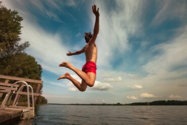 Ein Mann in roter Badehose springt mit ausgebreiteten von einem kleinen Steg in einen See.
