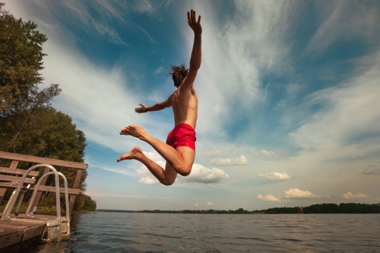 Ein Mann in roter Badehose springt mit ausgebreiteten von einem kleinen Steg in einen See.