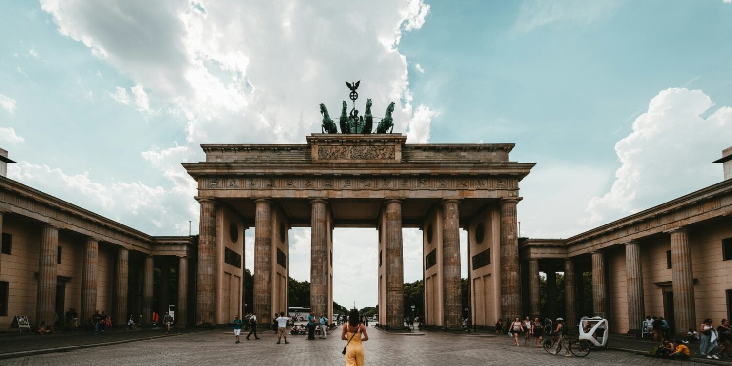 Eine Person in einem gelben Sommerkleid steht genau mittig vor dem Brandenburger Tor in Berlin.