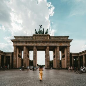 Eine Person in einem gelben Sommerkleid steht genau mittig vor dem Brandenburger Tor in Berlin.