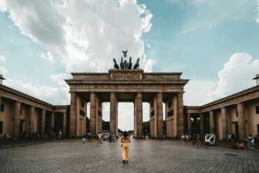 Eine Person in einem gelben Sommerkleid steht genau mittig vor dem Brandenburger Tor in Berlin.