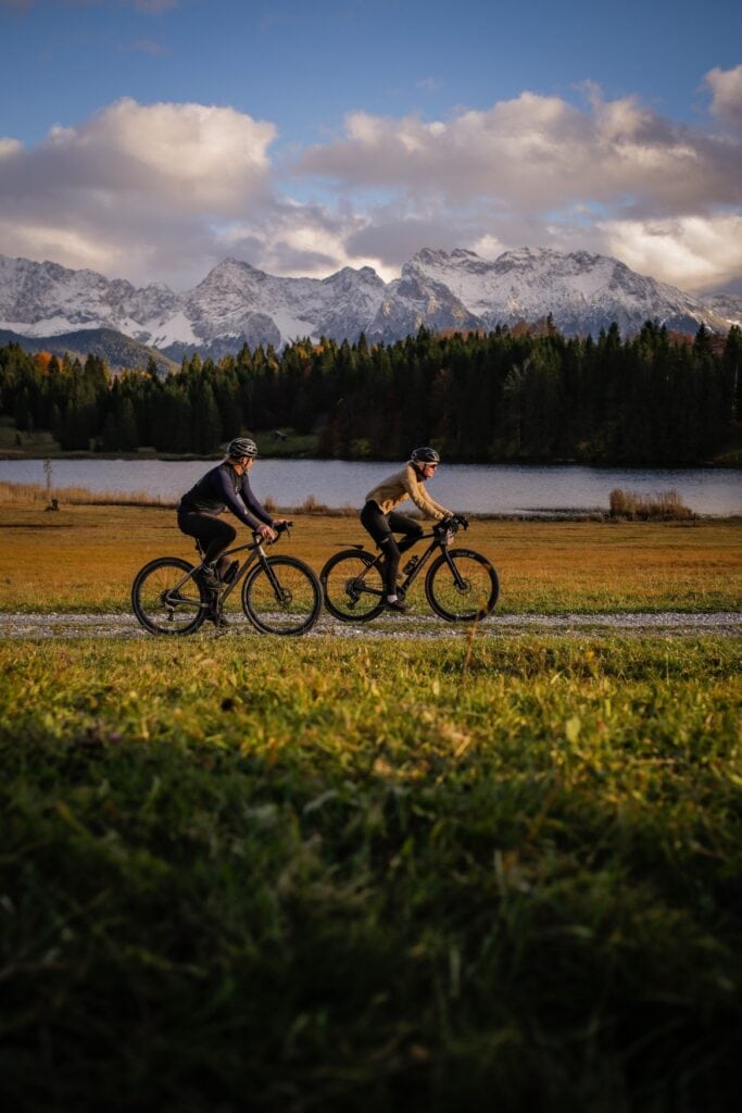 Zwei Personen fahren auf Gravelbikes auf einem Schotterweg an einem See entlang, im Hintergrund eine verschneite Bergkette der Alpen.