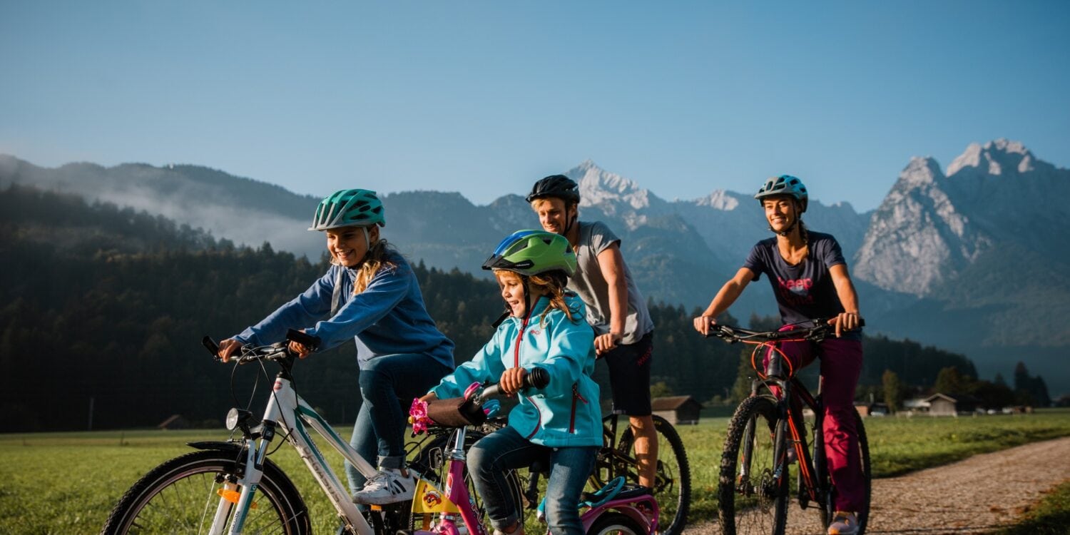 Zwei Kinder fahren mit ihren Eltern auf Fahrrädern einen Feldweg entlang, im Hintergrund sieht man einen Wald und eine Gipfelkette der Alpen bei Garmisch-Partenkirchen