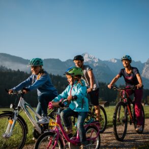 Zwei Kinder fahren mit ihren Eltern auf Fahrrädern einen Feldweg entlang, im Hintergrund sieht man einen Wald und eine Gipfelkette der Alpen bei Garmisch-Partenkirchen