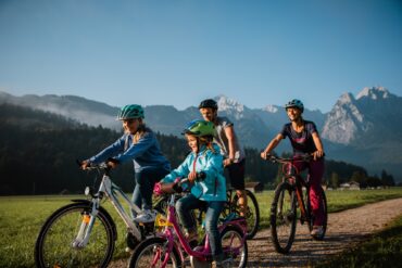Zwei Kinder fahren mit ihren Eltern auf Fahrrädern einen Feldweg entlang, im Hintergrund sieht man einen Wald und eine Gipfelkette der Alpen bei Garmisch-Partenkirchen