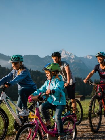 Zwei Kinder fahren mit ihren Eltern auf Fahrrädern einen Feldweg entlang, im Hintergrund sieht man einen Wald und eine Gipfelkette der Alpen bei Garmisch-Partenkirchen
