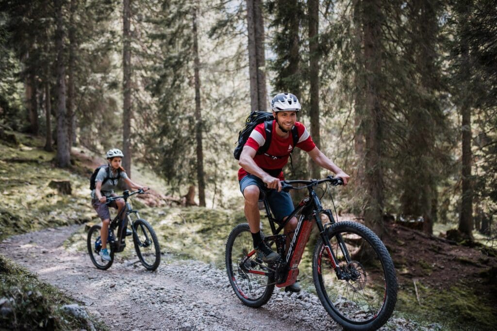 Ein junger Mann und ein Kind fahren mit elektrischen Mountainbikes eine Piste in einem Nadelwald in die Höhe.