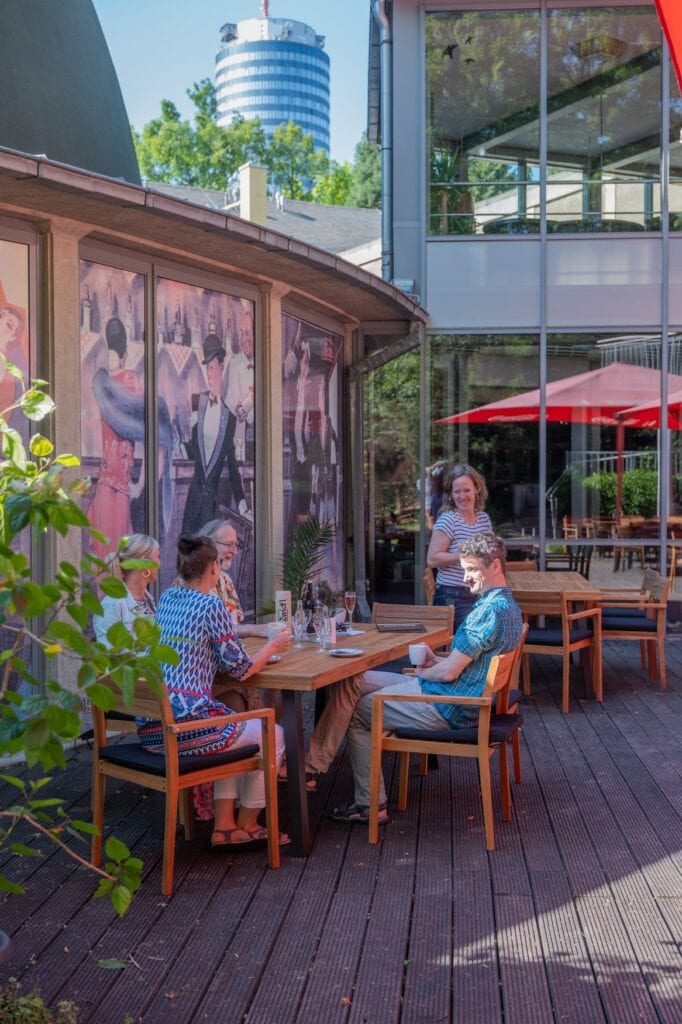 Besucher sitzen auf der Außenterrasse des Zeiss-Planetariums Jena, im Hintergrund der JenTower und das gläserne Planetariumsgebäude