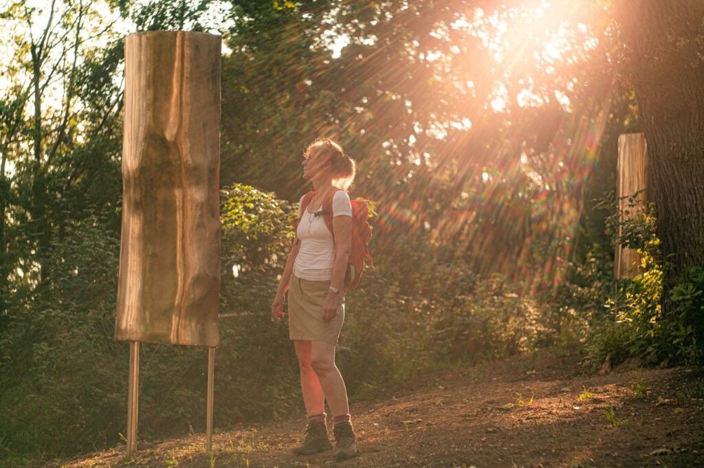 Wanderin mit Rucksack steht im Gegenlicht der Abendsonne zwischen zwei goldglänzenden Holzskulpturen auf dem Kunstweg bei Nebra. 