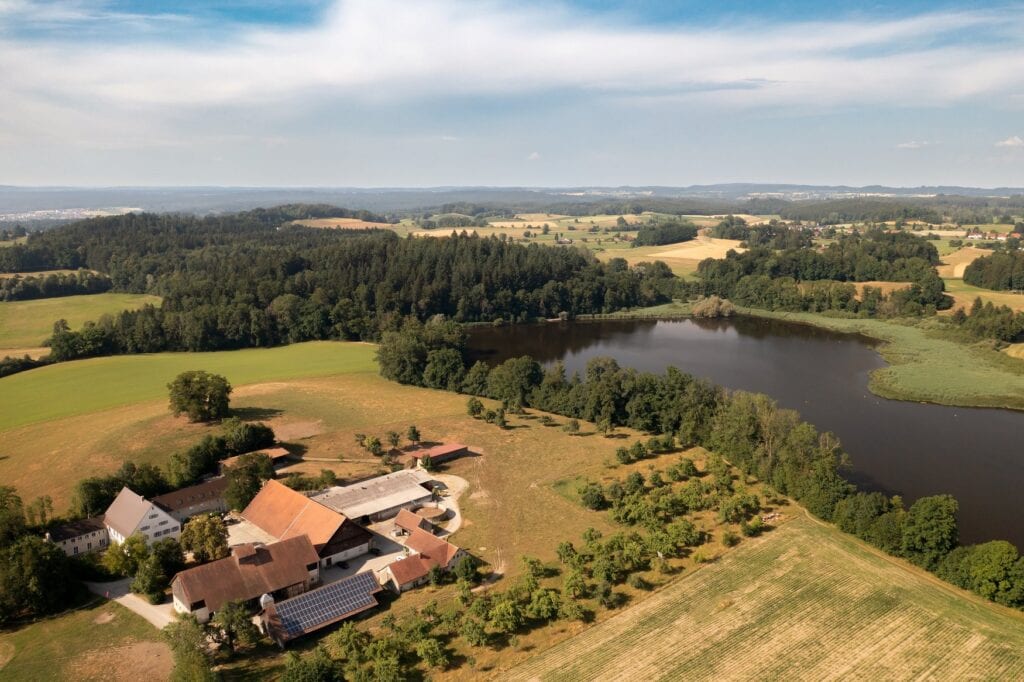 Luftaufnahme des Rösslerhofs mit seinen Gebäuden und Solaranlage, umgeben von Wiesen, Feldern und einem naturnahen See inmitten oberschwäbischer Landschaft. 