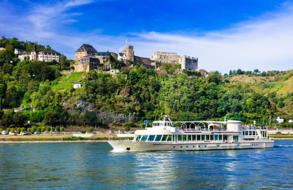 Der Rhein bei St. Goar, darauf ein weißes Fährschiff; am Ufer gegenüber ragt die Ruine der Burg Rheinfels mit angebundenem Schlosshotel Rheinfels aus einem kleinen Waldstück.