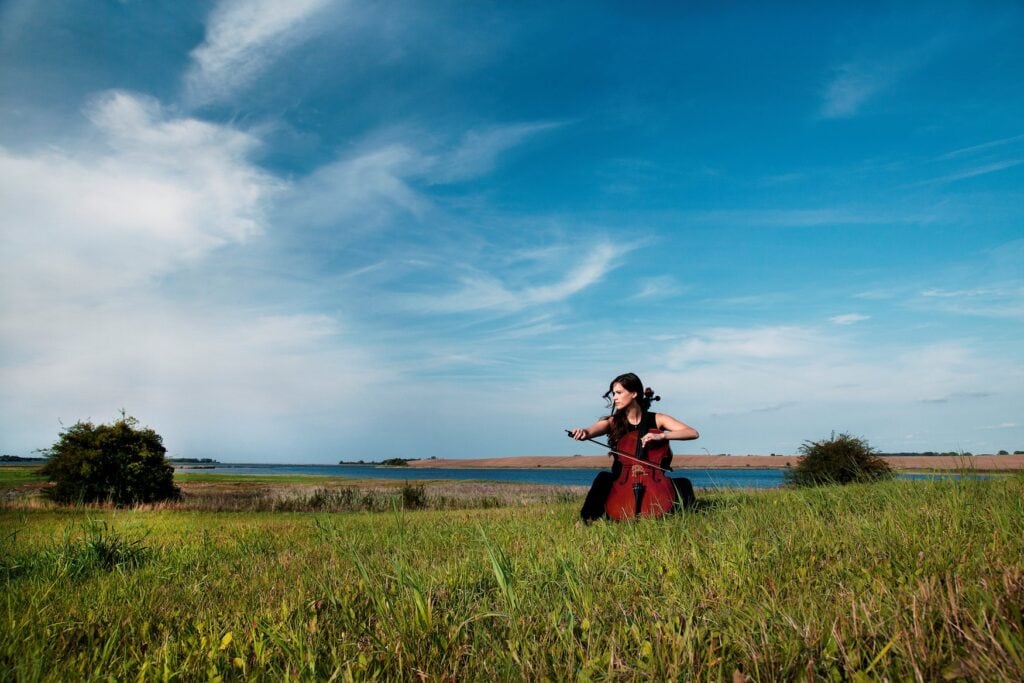 Eine Cellistin sitzt im hohen Gras einer Wiese am Wasser und spielt Violoncello. Über der weiten Boddenlandschaft steht ein blauer Himmel mit Schleierwolken. 