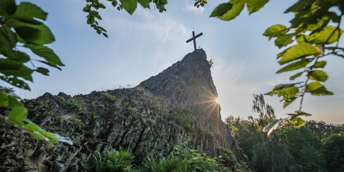 Blick auf den Druidenstein mit Gipfelkreuz, hinter dem sich die Sonne hervorstiehlt.