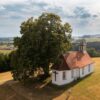 Aerial view of the Holy Cross Chapel on a hilltop surrounded by meadows, next to a large deciduous tree, with the rolling hills of Upper Swabia in the background.