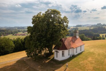 Aerial view of the Holy Cross Chapel on a hilltop surrounded by meadows, next to a large deciduous tree, with the rolling hills of Upper Swabia in the background.
