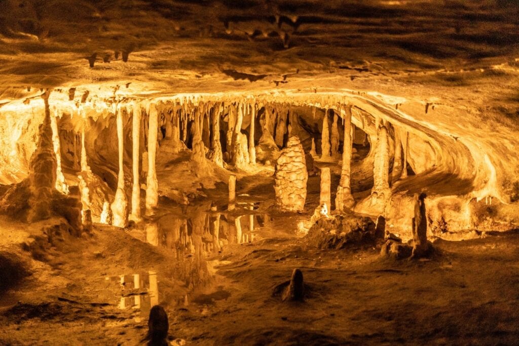 Stalaktiten hängen von der Decke der Hermannshöhle in Rübenland im Harz und spiegeln sich in einem Höhlensee, beleuchtet von warmem orangefarbenem Licht.