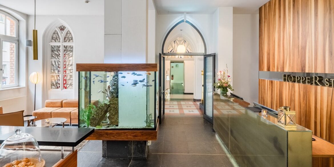 by des Hopper Hotels in einer umgebauten Kölner Kirche mit Empfangstresen, großem Aquarium als Raumteiler, gotischen Spitzbogenfenstern und Blick durch den historischen Kirchenbogen.