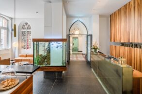 by des Hopper Hotels in einer umgebauten Kölner Kirche mit Empfangstresen, großem Aquarium als Raumteiler, gotischen Spitzbogenfenstern und Blick durch den historischen Kirchenbogen.
