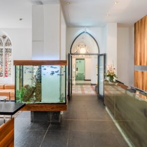 by des Hopper Hotels in einer umgebauten Kölner Kirche mit Empfangstresen, großem Aquarium als Raumteiler, gotischen Spitzbogenfenstern und Blick durch den historischen Kirchenbogen.