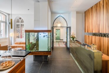 by des Hopper Hotels in einer umgebauten Kölner Kirche mit Empfangstresen, großem Aquarium als Raumteiler, gotischen Spitzbogenfenstern und Blick durch den historischen Kirchenbogen.