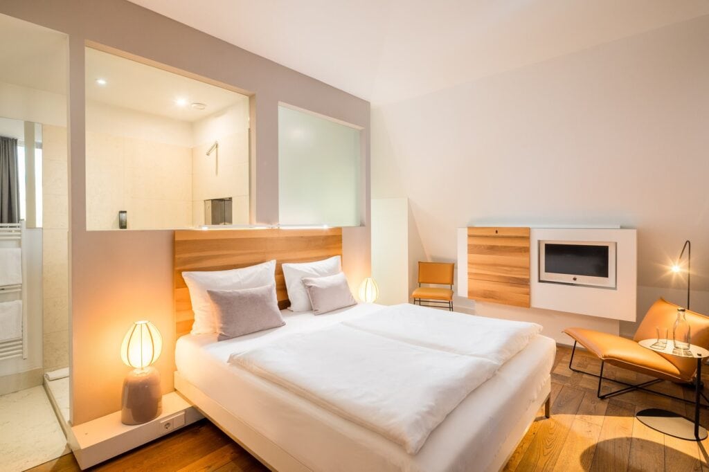 King-Size-Zimmer im Hopper Hotel Köln mit warmem Licht, Doppelbett mit Holzkopfteil, offenem Durchblick ins Badezimmer und cognacfarbenem Ledersessel.