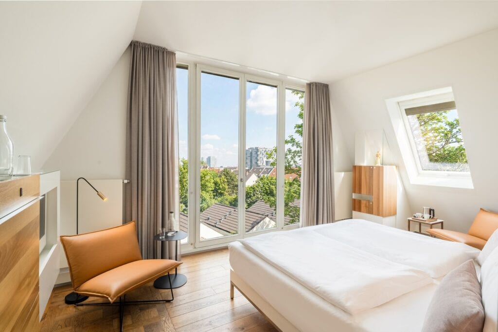 Helles King-Size-Zimmer im Hopper Hotel Köln mit Dachschräge, großem Panoramafenster mit Stadtblick auf Bäume und Dächer, cognacfarbenem Ledersessel und Holzparkett.