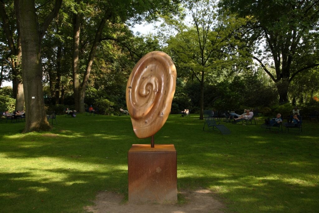 Im Luisenpark Mannheim steht auf einem rostbraunen Sockel eine große Holzskulptur in Form einer Schneckenspirale. Auf der sonnigen Wiese dahinter entspannen sich Menschen auf Liegen unter hohen Bäumen.