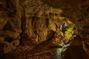 Zwei mit Stirnlampen ausgerüstete Personen klettern in der Tiefenhöhle Laichingen auf der Schwäbischen Alb über Felsblöcke durch einen weitläufigen Höhlengang.