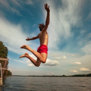 A man in red swim trunks jumps into a lake from a small dock with his arms outstretched.