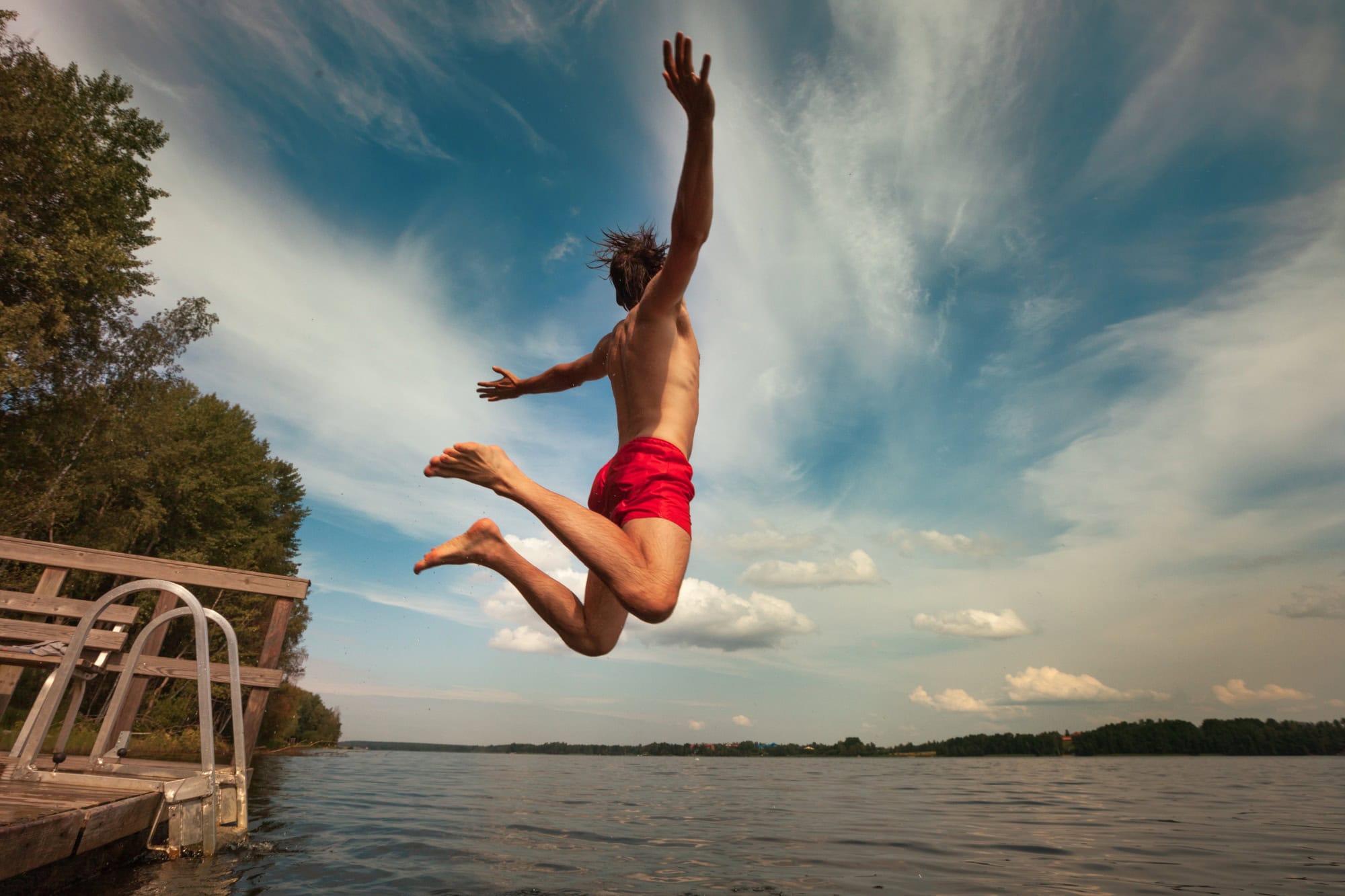 A man in red swim trunks jumps into a lake from a small dock with his arms outstretched.