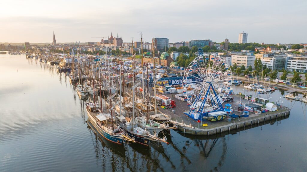 Luftaufnahme des Rostocker Stadthafens während der Hanse Sail. Am Kai liegen zahlreiche historische Segelschiffe. Rechts dreht sich ein großes Riesenrad, dahinter erstreckt sich die Stadtsilhouette von Rostock.