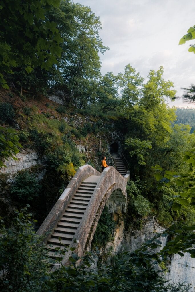 Eine steinerne Bogenbrücke mit Treppenstufen führt durch eine bewaldete Felsschlucht im Park Inzigkofen. Auf der Brücke sitzt eine Person in oranger Jacke und blickt in die Landschaft.