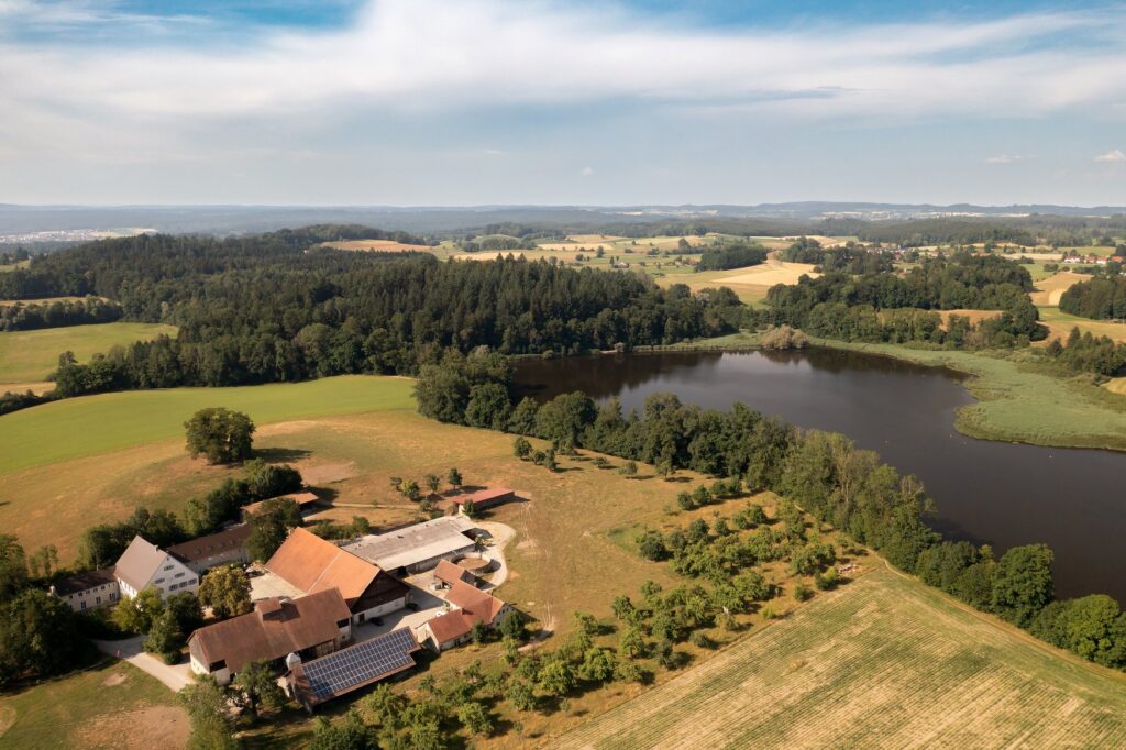 Aerial view of the Rösslerhof with its buildings and solar power system, surrounded by meadows, fields, and a near-natural lake in the heart of the Upper Swabian countryside.