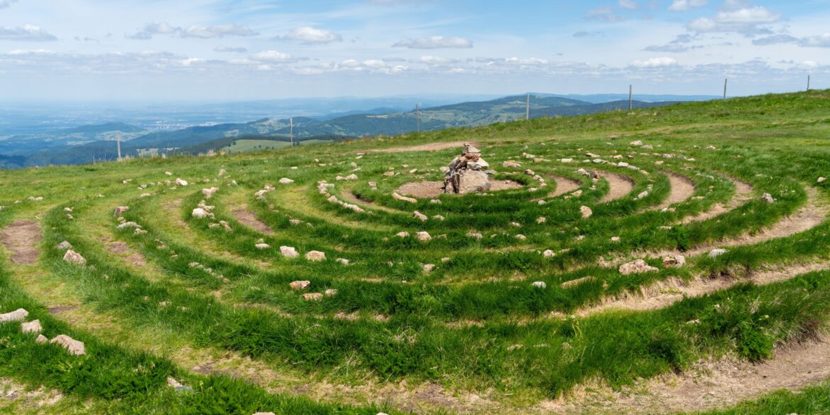 Auf einer Bergwiese am Belchen im Schwarzwald liegt ein aus Steinen gelegtes Spirallabyrinth mit einem Steinhaufen in der Mitte. Im Hintergrund öffnet sich der weite Blick über bewaldete Höhenzüge.