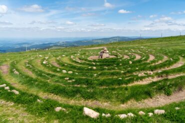 Auf einer Bergwiese am Belchen im Schwarzwald liegt ein aus Steinen gelegtes Spirallabyrinth mit einem Steinhaufen in der Mitte. Im Hintergrund öffnet sich der weite Blick über bewaldete Höhenzüge.