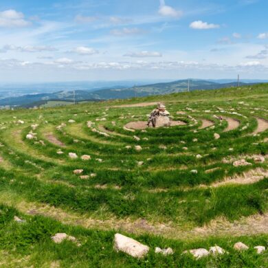 Auf einer Bergwiese am Belchen im Schwarzwald liegt ein aus Steinen gelegtes Spirallabyrinth mit einem Steinhaufen in der Mitte. Im Hintergrund öffnet sich der weite Blick über bewaldete Höhenzüge.