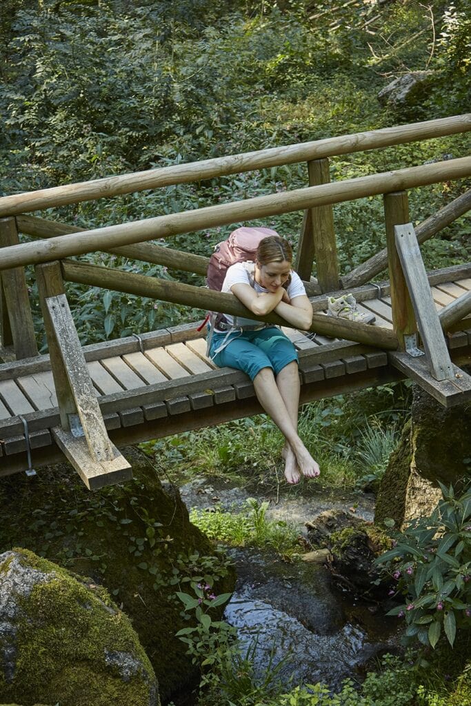Eine Wanderin mit Rucksack sitzt barfuß auf einer hölzernen Fußgängerbrücke über einem kleinen Waldbach und stützt die Arme auf dem Geländer ab. Ihre Wanderschuhe stehen neben ihr, ringsum wächst dichtes Grün.