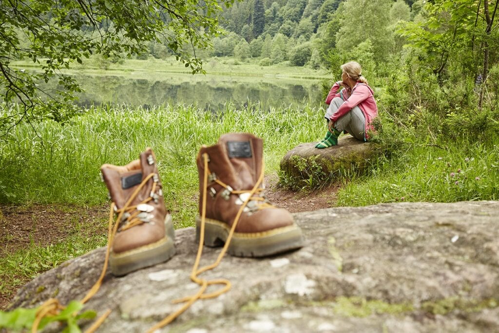 Eine Wanderin sitzt auf einem Felsen am Ufer eines ruhigen Waldsees und blickt über das Wasser. Im Vordergrund stehen ihre abgestellten braunen Wanderschuhe auf einem Stein.