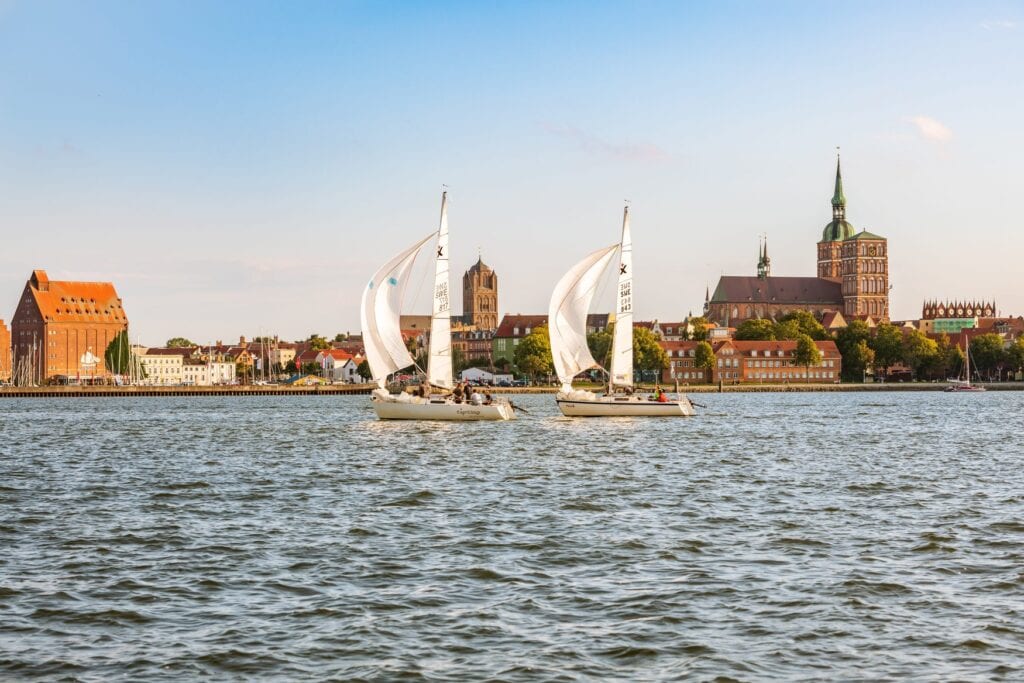 Zwei Segelboote kreuzen bei der Stralsunder Mittwochsregatta auf dem Wasser vor der Altstadtsilhouette. Im Hintergrund ragt die Nikolaikirche mit ihrem grünen Turm auf.