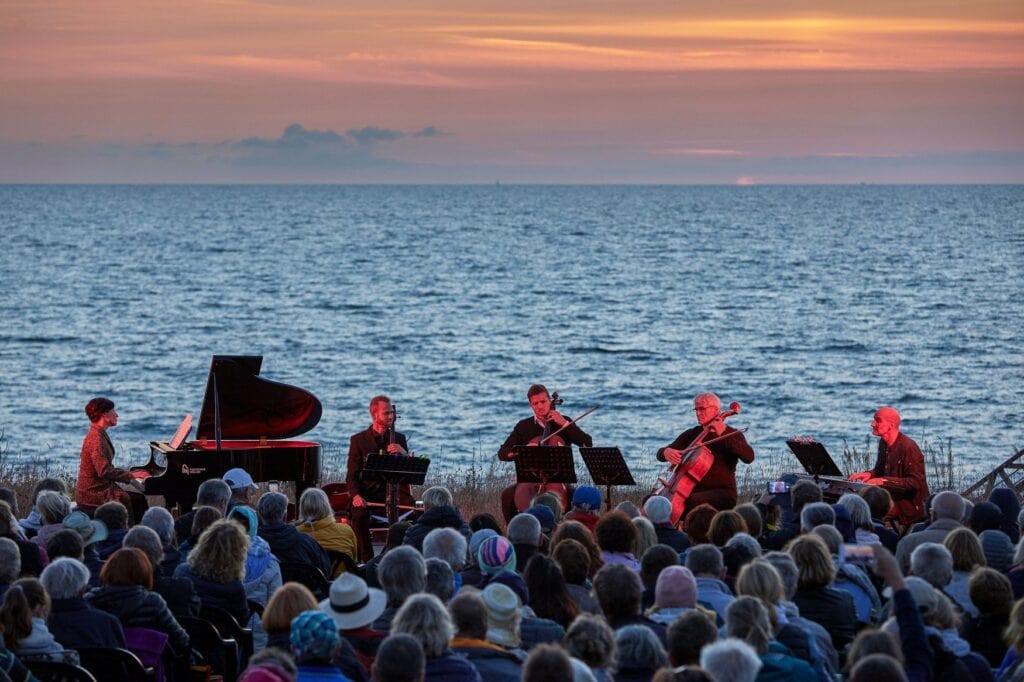 Abendkonzert der Naturklänge am Meer im Ostseebad Ahrenshoop. Ein Ensemble mit Flügel und Violoncelli spielt vor zahlreichen Zuhörern, während sich der Himmel über der Ostsee orange färbt.
