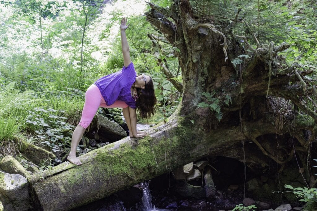 Eine Frau in violettem Oberteil und rosa Hose macht barfuß auf einem bemoosten, umgestürzten Baumstamm über einem Waldbach eine Yoga-Übung im gedrehten Dreieck. Ringsum wächst dichter grüner Farn- und Laubwald.