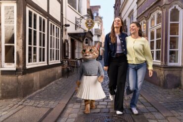A costumed cat character in a jumper and pleated skirt walking with two laughing women along a cobblestone lane in the Schnoor quarter.