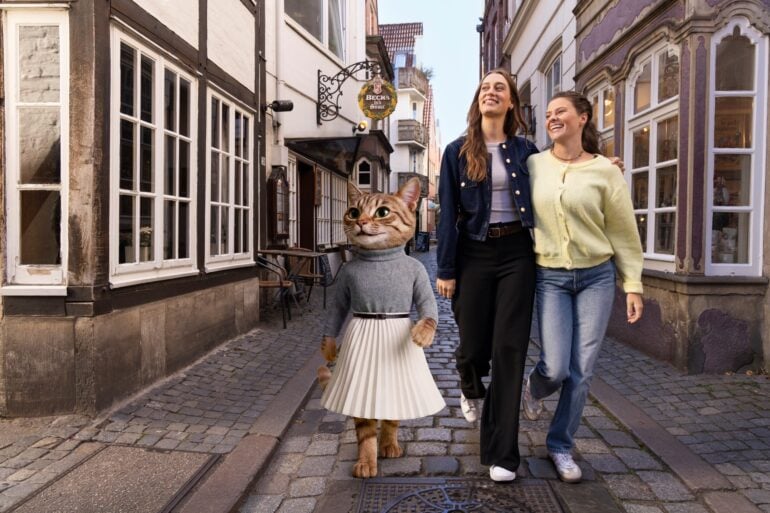 A costumed cat character in a jumper and pleated skirt walking with two laughing women along a cobblestone lane in the Schnoor quarter.