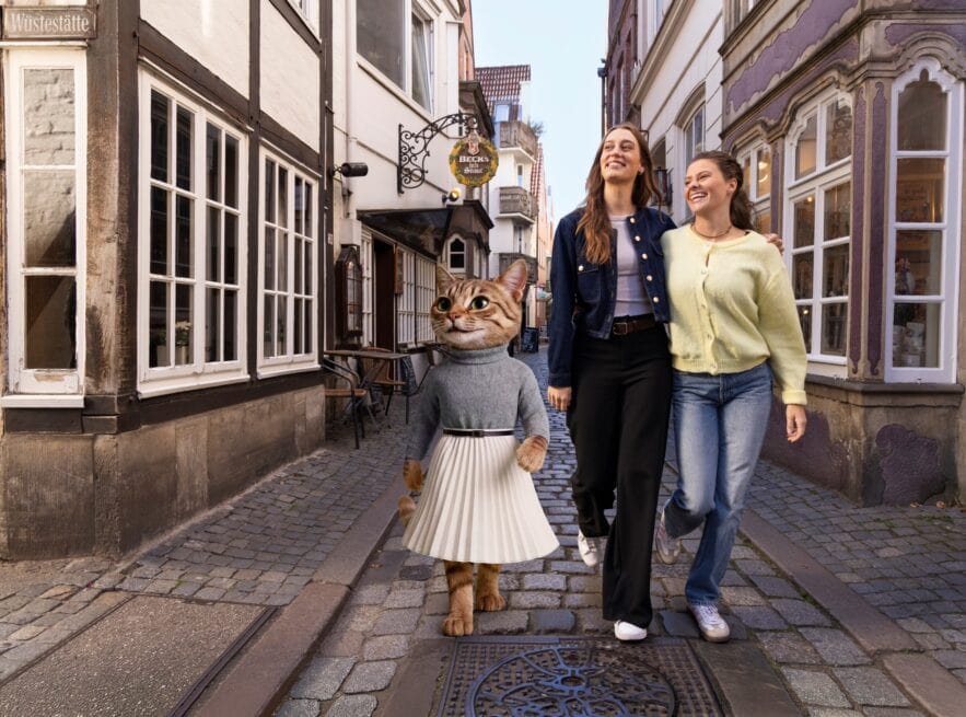 A costumed cat character in a jumper and pleated skirt walking with two laughing women along a cobblestone lane in the Schnoor quarter.