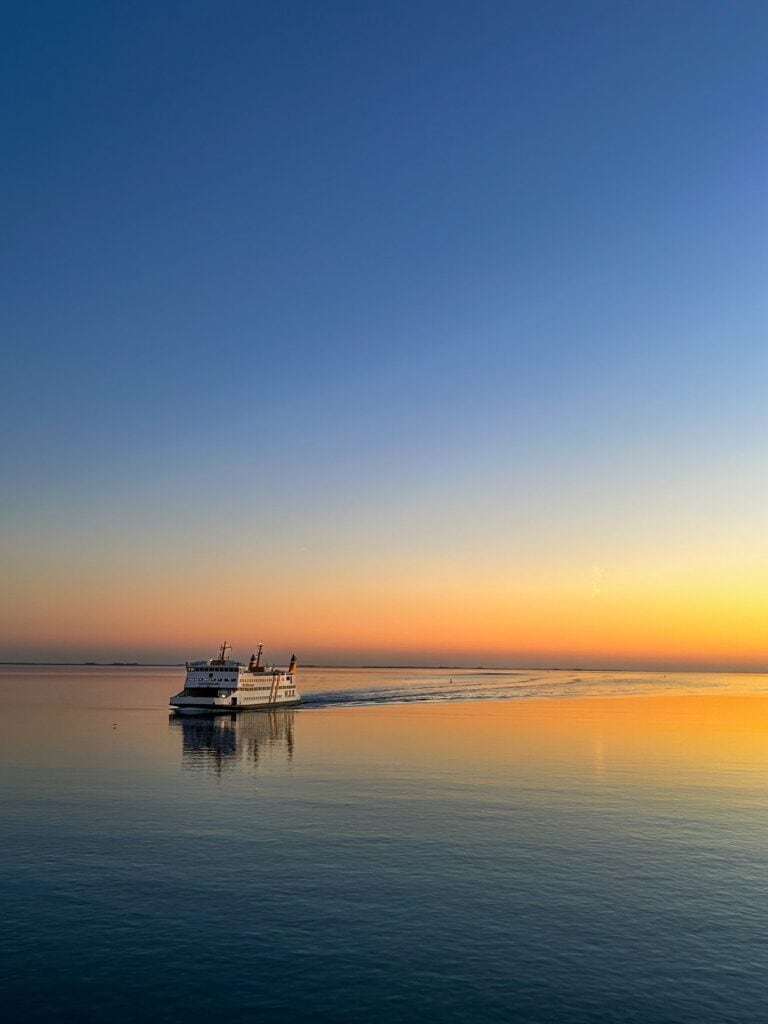 Fähre der W.D.R. fährt bei Sonnenuntergang über das ruhige Wattenmeer, der Himmel geht von Blau in Orange über.